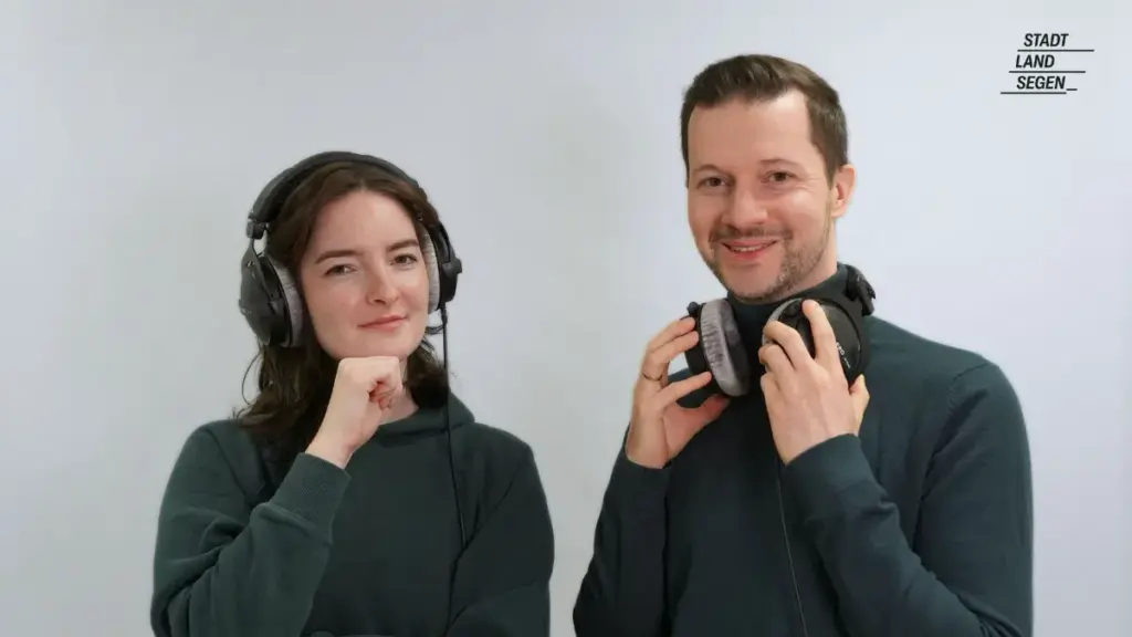 Das Bild zeigt zwei Menschen, die freundlich in die Kamera lächeln. Beide tragen einen Kopfhörer. Die Linke Person, Miriam, hat den Kopfhörer auf beiden Ohren aufgesetzt. Die rechte Person, Christian, trägt den Kopfhörer lässig um den Hals.